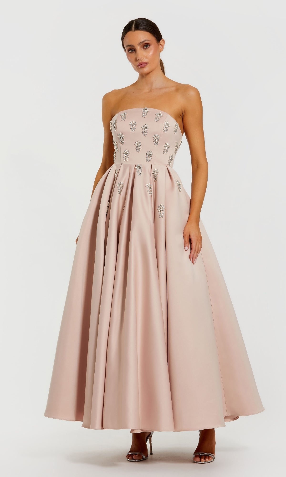 Woman wearing a strapless blush pink dress with crystal embellishments stranding in front of a white background